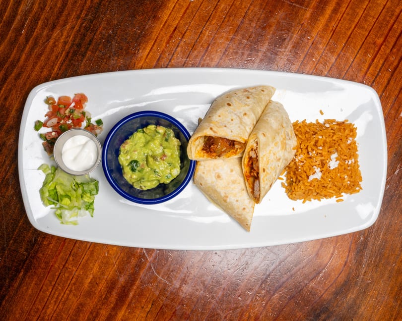 Two burritos with rice, guacamole, sour cream, pico de gallo, and lettuce on a white plate.