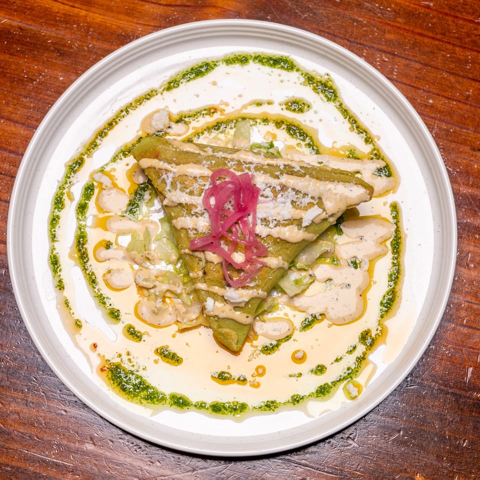 Green folded crepe topped with pickled red onions, sauce, and cheese on a white plate.