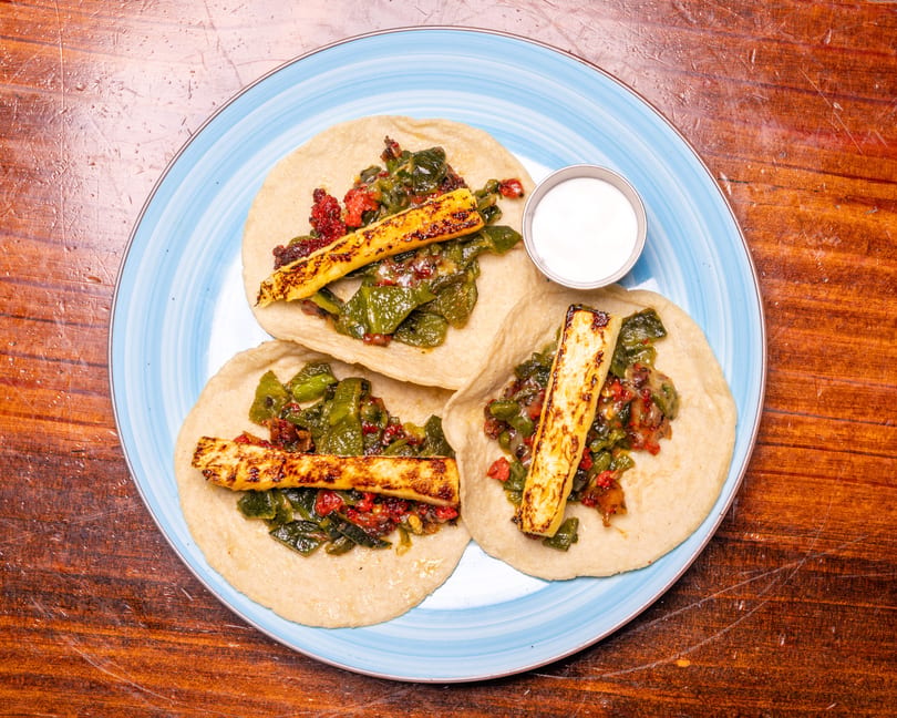 Three tacos with green and red filling, grilled cheese sticks, and a side of white sauce.