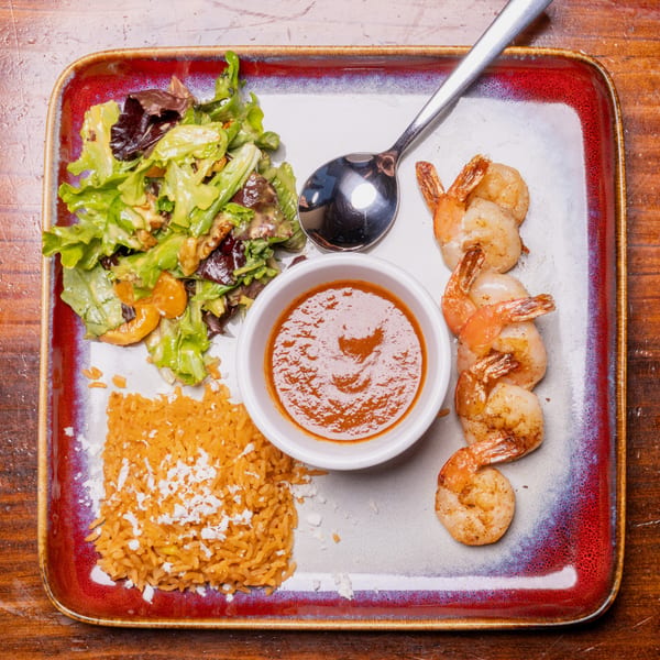 Grilled shrimp, rice, salad, and a bowl of sauce on a rectangular plate.