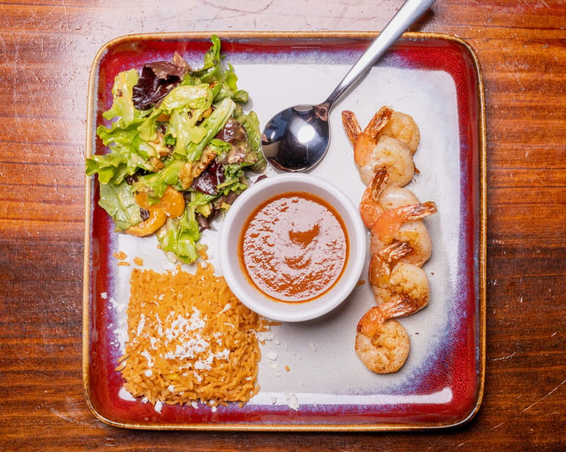Grilled shrimp, rice, salad, and a bowl of sauce on a rectangular plate.