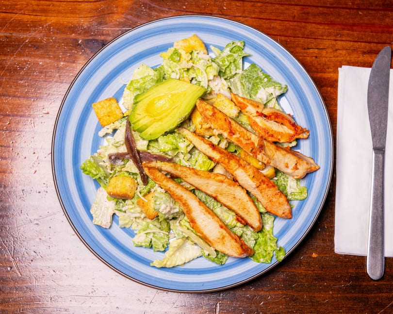 Caesar salad with grilled chicken, avocado, anchovies, and croutons on a blue striped plate.