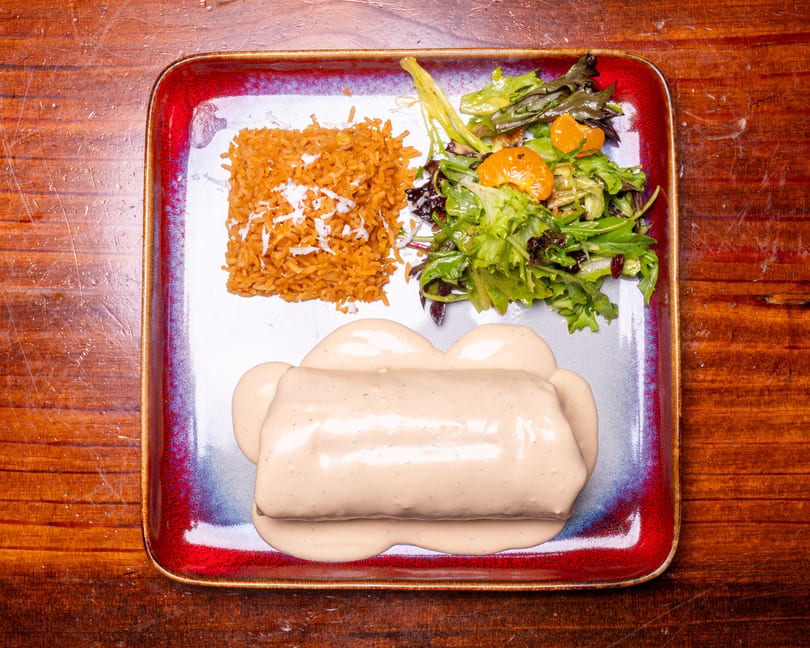 A burrito covered in creamy sauce, with a side of rice and a green salad with mandarin oranges.