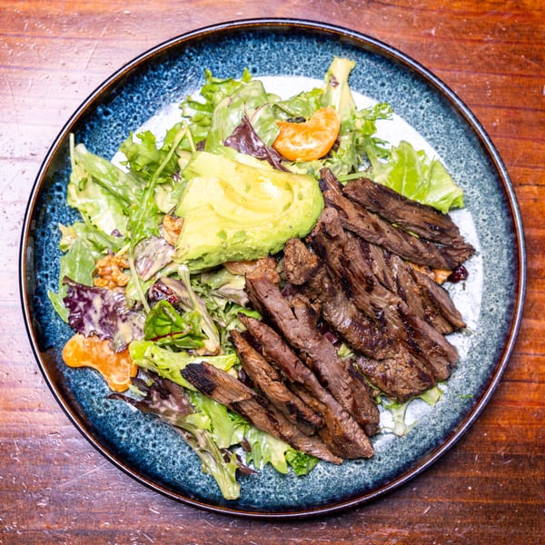 Grilled steak salad with avocado, mixed greens, mandarin oranges, and walnuts on a blue plate.