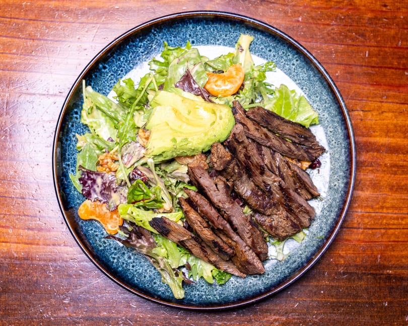 Grilled steak salad with avocado, mixed greens, mandarin oranges, and walnuts on a blue plate.