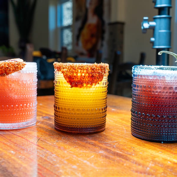 Three colorful cocktails in textured glasses, two with chili-lime rims, on a wooden table.