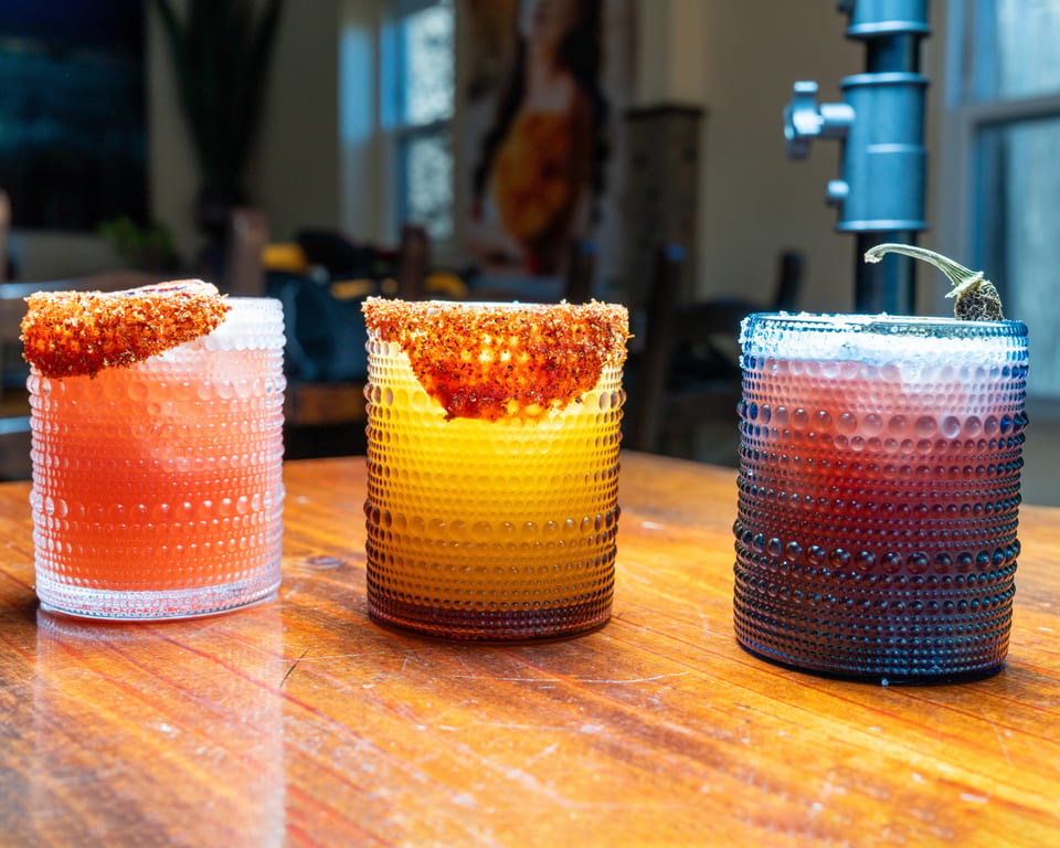 Three colorful cocktails in textured glasses, two with chili-lime rims, on a wooden table.