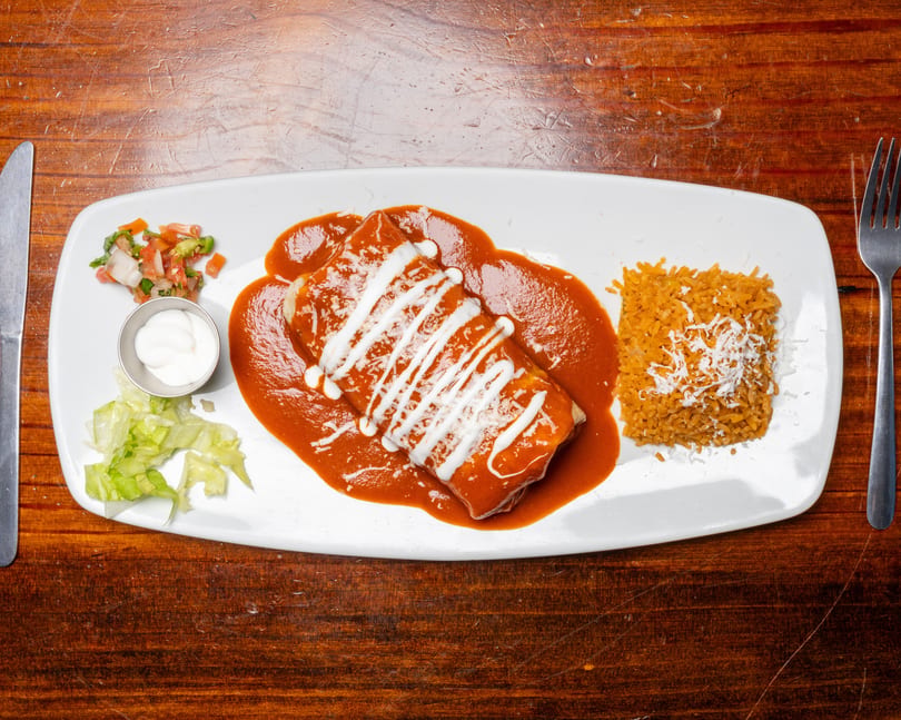 A burrito covered in red sauce and sour cream, with rice, salsa, and lettuce on a white plate.