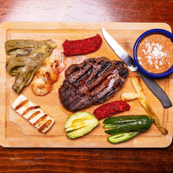 Grilled steak, shrimp, vegetables, and beans on a wooden cutting board with a knife.
