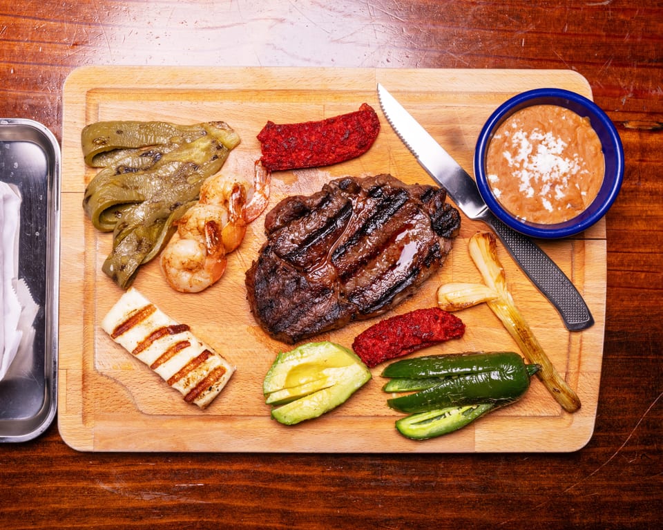Grilled steak, shrimp, vegetables, and beans on a wooden cutting board with a knife.