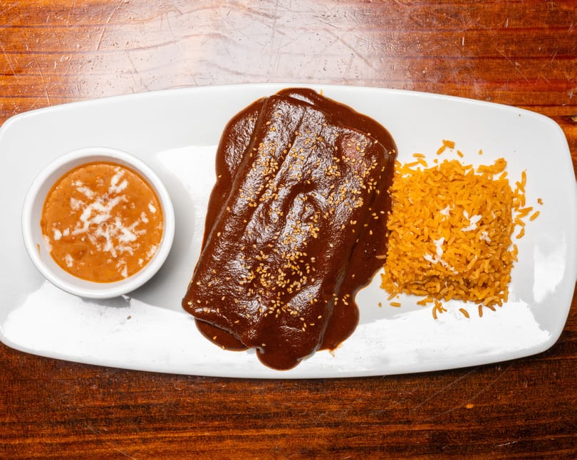 Enchiladas with mole sauce and sesame seeds, served with rice and a side of beans.