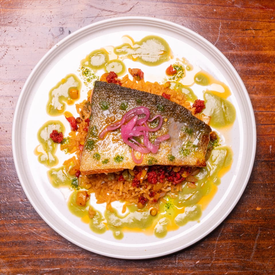 Crispy-skinned fish fillet with pickled onions, rice, red relish, and green sauce on a white plate.