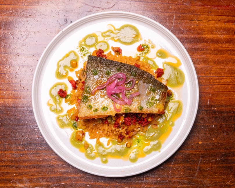 Crispy-skinned fish fillet with pickled onions, rice, red relish, and green sauce on a white plate.