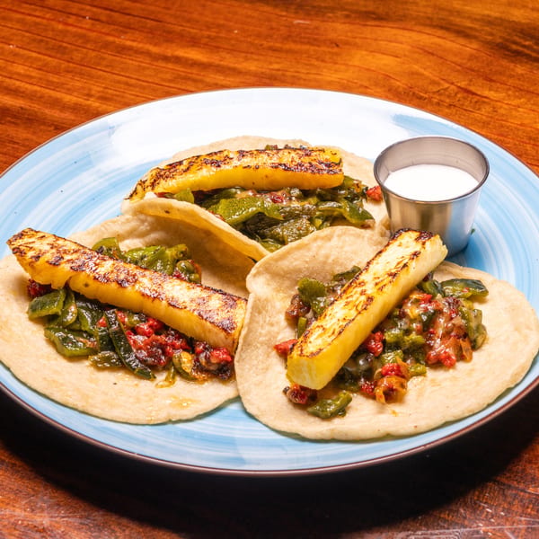 Three tacos with grilled cheese, green peppers, and red sauce, served with a side of white sauce.