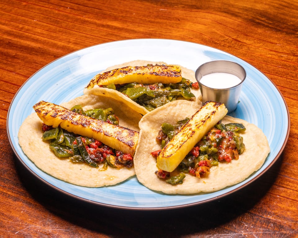 Three tacos with grilled cheese, green peppers, and red sauce, served with a side of white sauce.