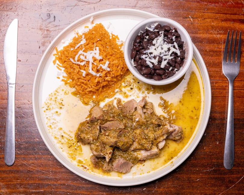 Pork in green sauce with rice and black beans, topped with cheese, on a white plate.
